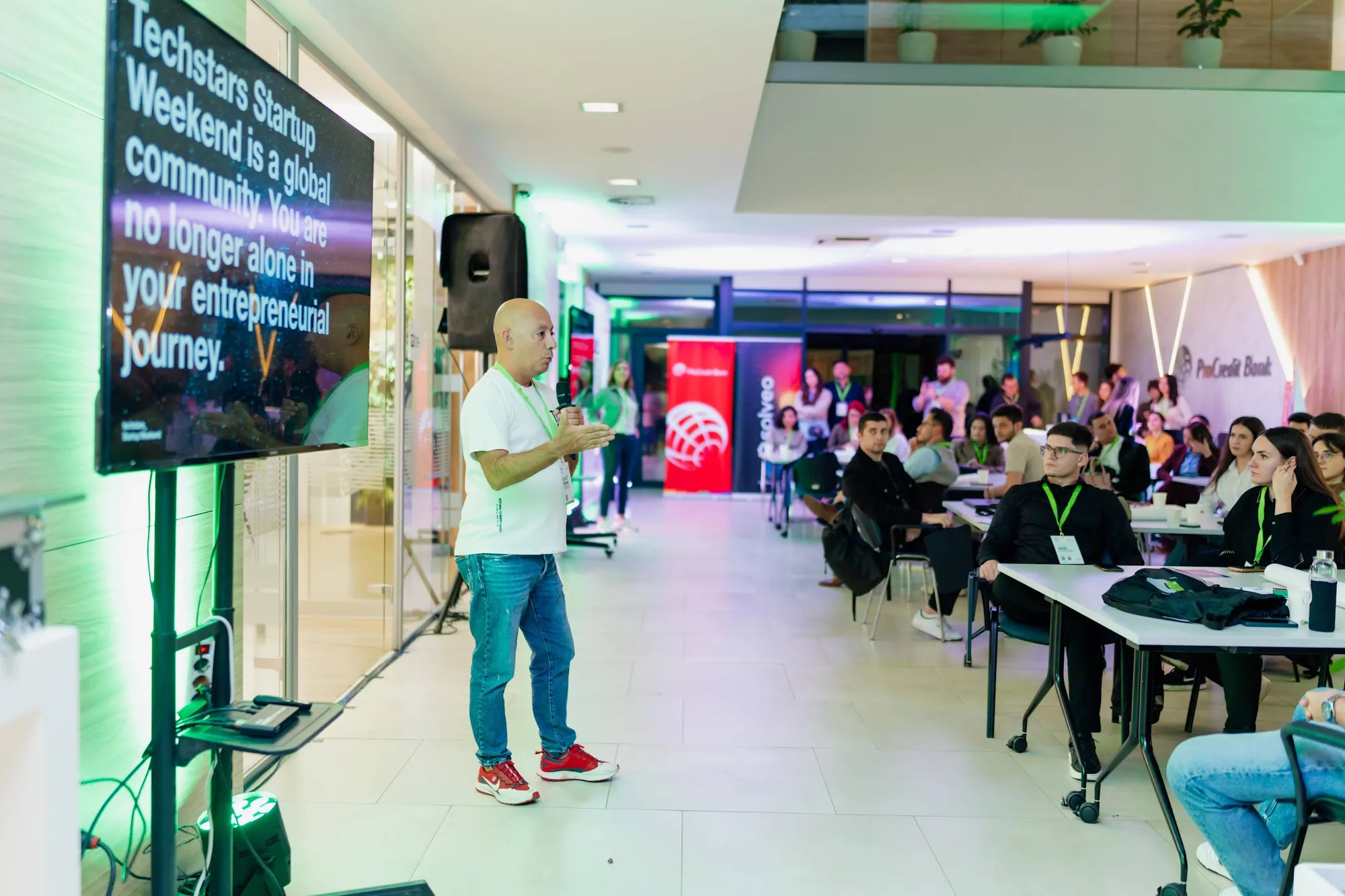 The facilitator opening a past Startup Weekend Skopje in the venue's atrium, with the audience at tables and a screen reading “Startup Weekend is a global community” beside him